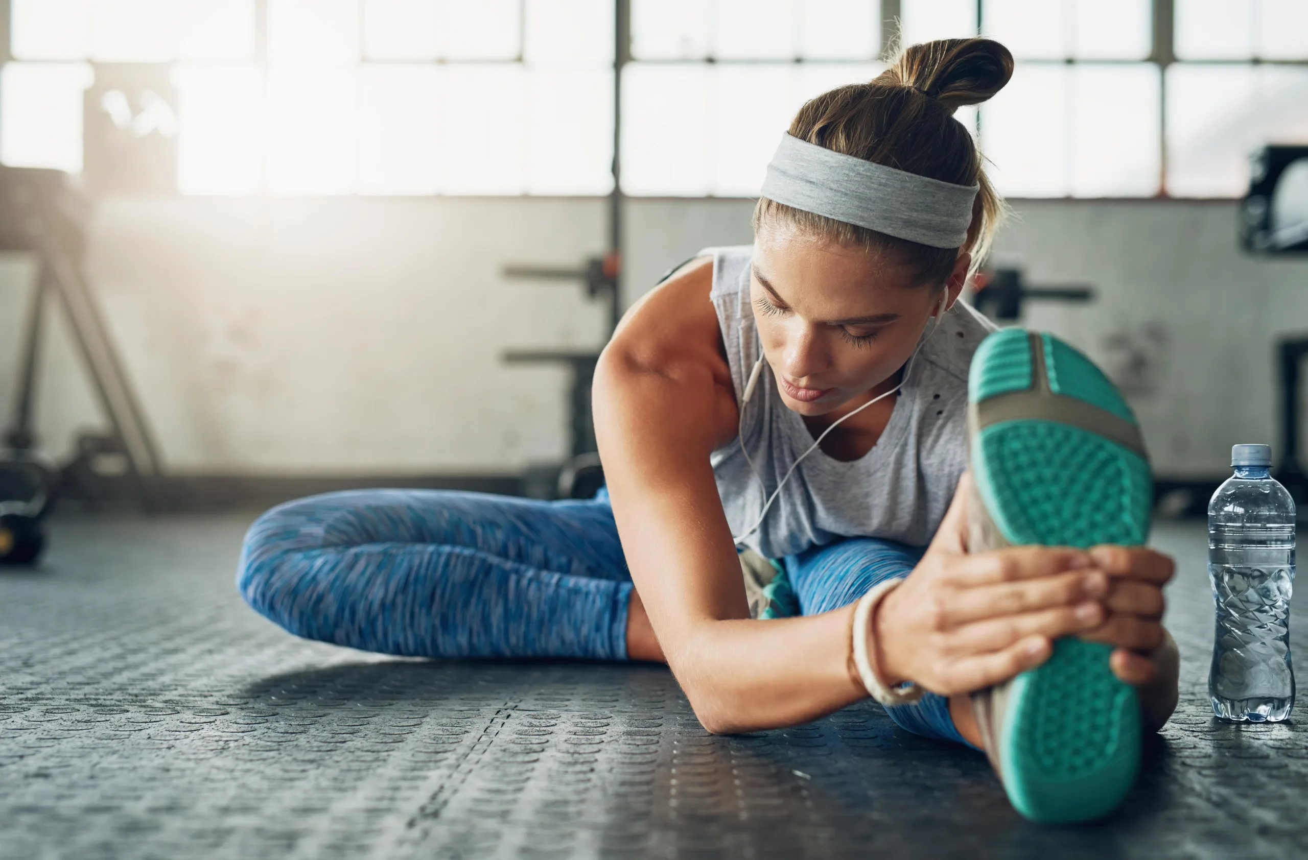 Woman stretching her hamstrings on the gym floor. She has on a grey headband, wire earphones, blue pants and blue soles of her shoes. There is a water bottle beside her, she seems focused and is looking down.