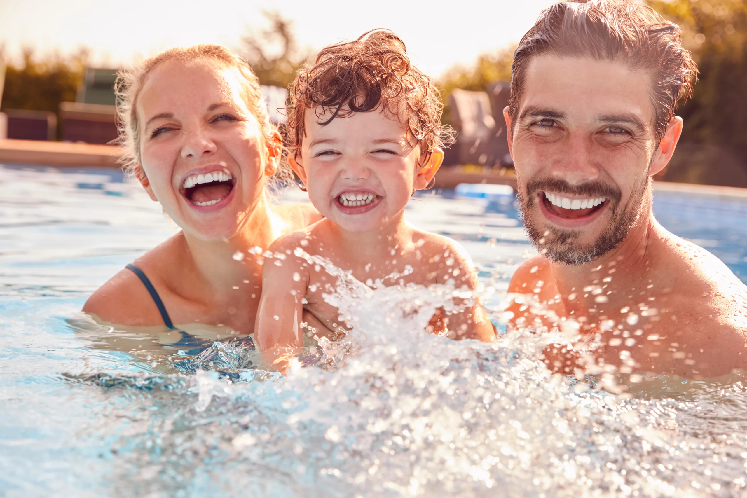 Family splashing and laughing in the pool. Mom on the left with fair skin and hair slicked back laughing holding toddler son smiling and splashing in the water with dad on the right who is fair skinned and has a laughing smile with a short shaggy beard also holding him all looking at the camera in a candid moment in the pool.